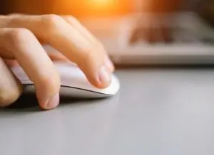 A close-up of a hand using a white computer mouse on a gray surface, with part of a laptop visible in the background and warm sunlight shining from above.