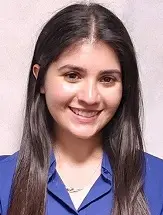 A young woman with long, straight dark hair is smiling. She is wearing a blue collared shirt and a necklace, standing in front of a plain, light-colored background.