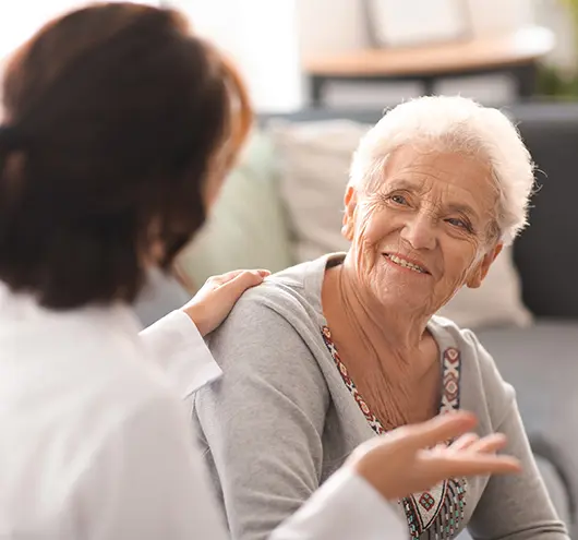 An elderly woman with short gray hair smiles warmly while sitting on a couch, engaged in conversation with a younger woman who has her hand on the elderly womans shoulder.
