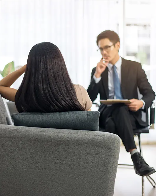A woman with long dark hair sits on a gray chair, facing a man in a suit who holds a clipboard, suggesting a professional conversation or counseling session in a bright office setting.