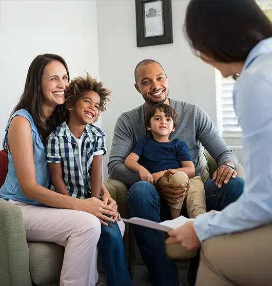 A smiling family with two children sits together on a couch, talking to a professional woman holding a clipboard in a bright, comfortable living room.
