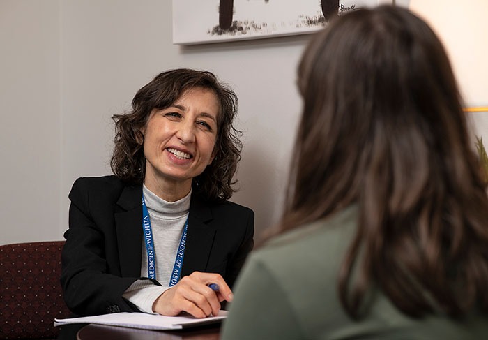 A woman in a blazer and lanyard smiles while talking to another person across a desk, holding a pen and notepad. The background includes a white wall and part of a lamp.
